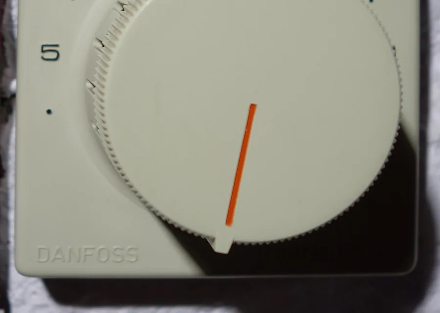 A wall mounted room thermostat with a numbered dial and an orange pointer showing the setting.