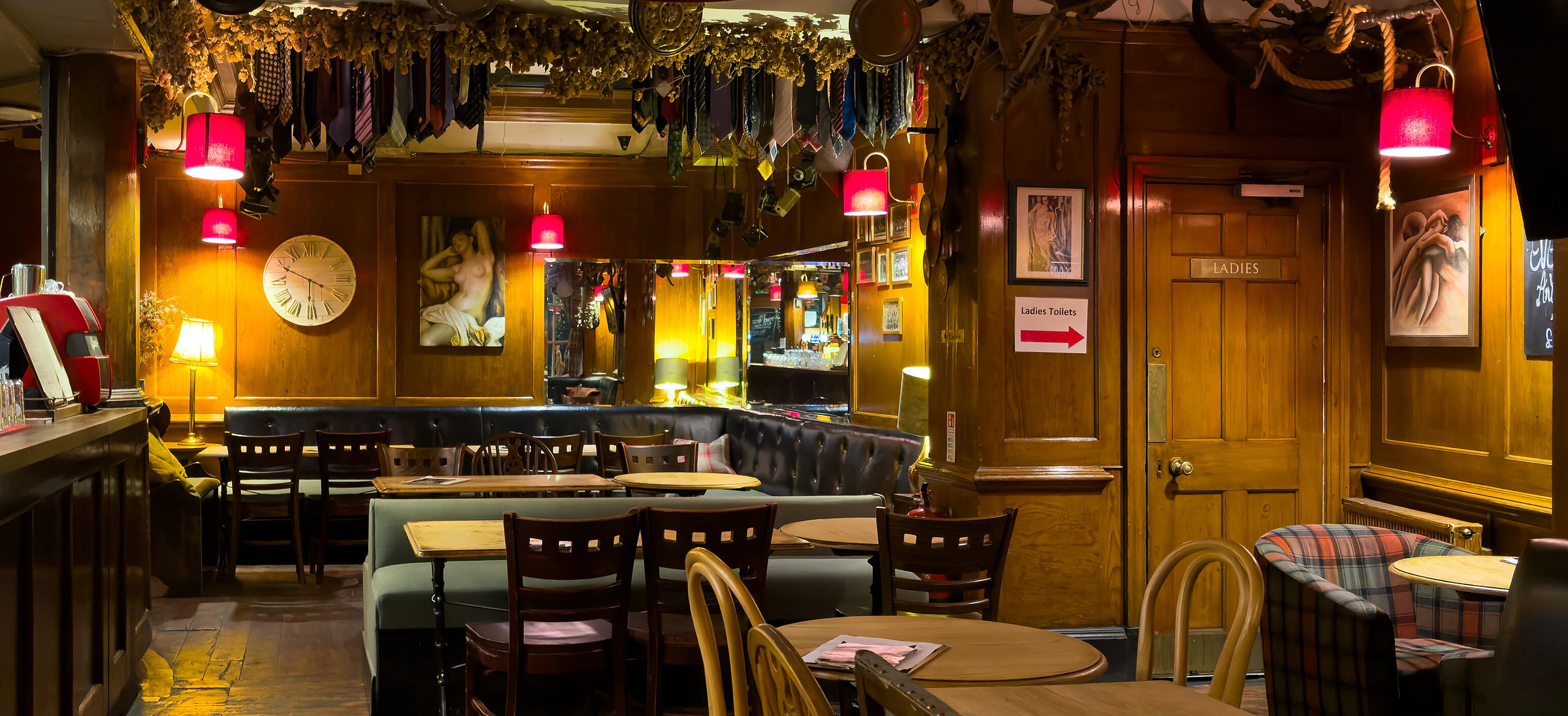 The lounge: hop bines and old ties strung along the beams, red-shaded lamps over panelled walls, a wall clock, and leather banquettes and wooden tables running back into the room.